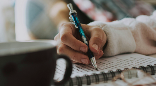 Une personne écrit dans un cahier à spirale avec un stylo bleu, concentrée sur la conjugaison et les participes passés. Une tasse noire l'accompagne.